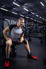 Fototapeta na wymiar A athlete with blond hair shakes biceps in dumbbells in the gym.