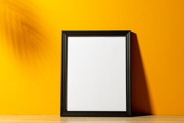 Black empty frame with copy space on table against yellow wall and shadow of leaves