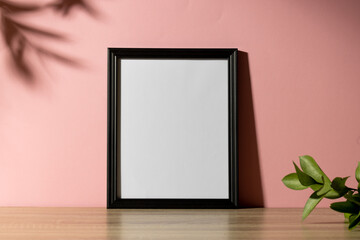 Fototapeta na wymiar Black empty frame with copy space with plant on table against pink wall and shadow of leaves