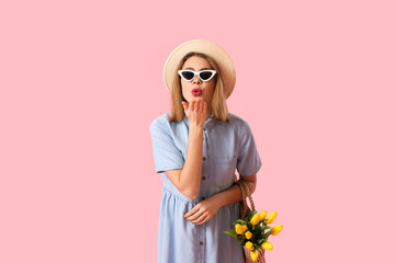 Young woman in sunglasses with bag of tulips blowing kiss on pink background