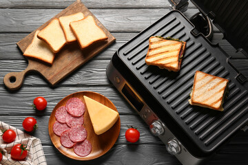 Composition with modern electric grill, delicious sandwiches and ingredients on black wooden background