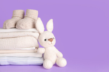 Stack of baby clothes, socks and knitted toy on lilac background