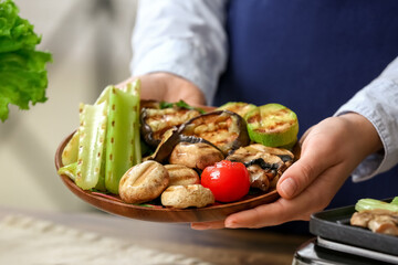 Woman holding plate with tasty grilled vegetables at table in kitchen, closeup