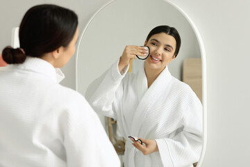 Young woman removing makeup with cotton pad near mirror in bathroom