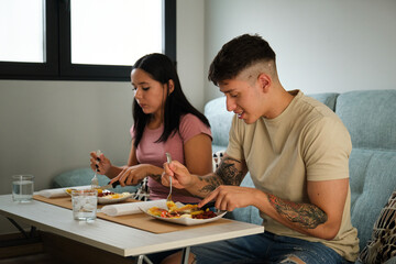 Two young hispanic people eating traditional llapingachos sitting on the sofa.