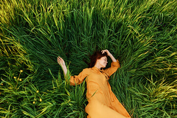 an elegant woman lies in the tall green grass in a long summer orange dress and with her arms...