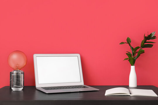 Workplace with laptop, lamp and notebook near red wall