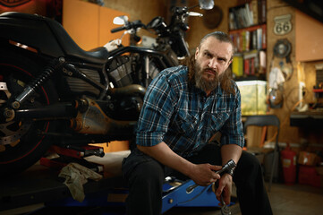 Obraz na płótnie Canvas Portrait of mature bearded biker looking at camera sitting in his own garage