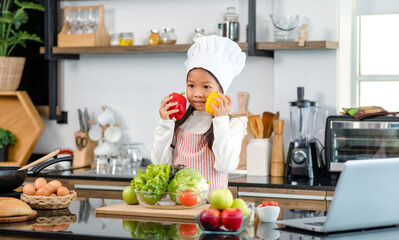 Millennial Asian young little cute girl chef daughter with white tall cook hat and apron standing smiling holding wooden spoon and fork posing ready for cooking vegetables at counter in home kitchen