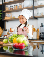 Millennial Asian young little cute girl chef daughter with white tall cook hat and apron standing smiling holding wooden spoon and fork posing ready for cooking vegetables at counter in home kitchen