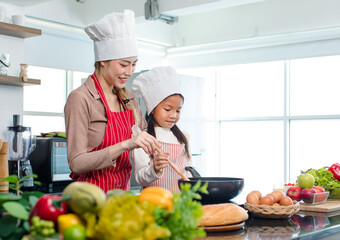 Asian young little cute girl chef daughter wears white tall cook hat and apron standing smiling posing together with mother while cooking food with pan in home kitchen.