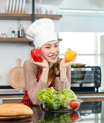 Asian young female chef wears white tall cook hat and apron smiling posing holding red yellow sweet peppers ready to cooking food with pan at counter with bread vegetables ingredients in home kitchen