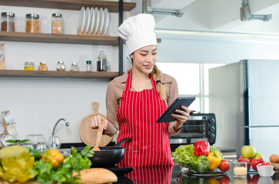 Asian young female chef housewife wears white tall cook hat and apron learning cooking homemade food meal online via touchscreen tablet computer while using spatula frying eggs in pan at home kitchen