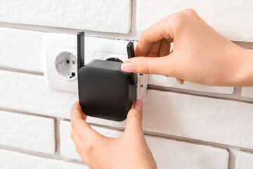 Woman plugging black WiFi repeater in electric socket on white brick wall