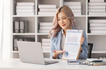 A young Asian businesswoman shows documents with graphs with a pen, holds in his hand smiles. Sitting at a desk with a laptop in a modern office.