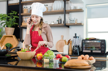 Asian young female chef housewife wears white tall cook hat and apron standing smiling on smartphone call holding wooden spoon fork spatula mixing delicious vegetable salad in bowl at home kitchen