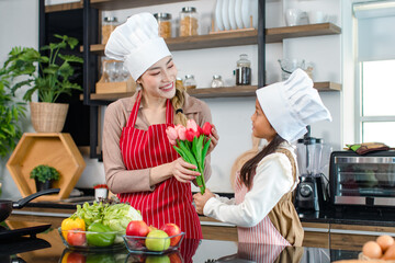 Asian young cheerful female chef housewife mother and little girl daughter wears white tall cook hat and apron standing smiling holding tulip flower bouquet together in kitchen preparing food meal