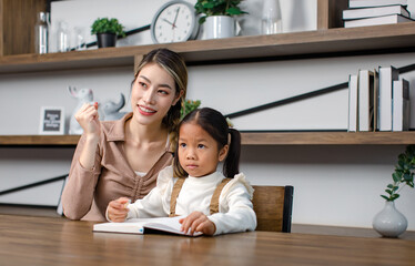 Asian young female housewife mother tutor teacher sitting smiling on table in living room at home teaching little cute kindergarten preschool girl daughter writing on book doing homework after school