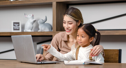 Asian young female housewife mother tutor teacher sitting smiling on table in living room using notebook computer pointing teaching little cute kindergarten preschool girl daughter doing homework
