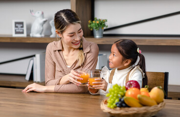 Obraz na płótnie Canvas Asian young female housewife mother sitting smiling on table near bookshelf in living room at home with little girl kid daughter holding healthy organic orange juice glasses cheers drinking together