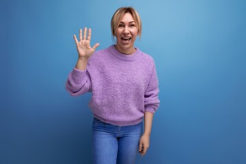 cute blond young woman in purple hoodie shows her palm on a blue background with copy space