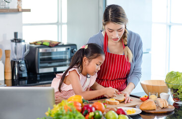 Asian young female chef mother wears apron teaching little cute girl daughter cooking preparing making delicious sandwiches with vegetables sliced bread egg butter and ingredients in home kitchen