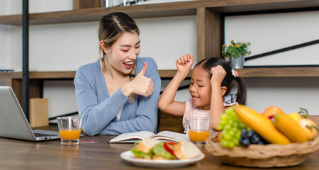Asian young female mother and little cute girl daughter sitting smiling learning studying doing homework online via laptop notebook computer drinking orange juice eating sandwich together at home