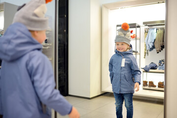 Cute little boy is trying on a new coat and hat in front of a mirror in a store or shopping mall....
