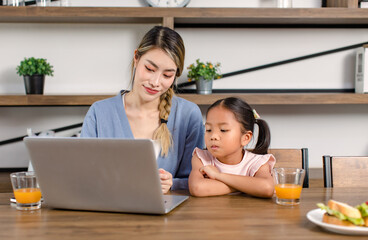 Asian young cheerful female mother and little cute girl daughter sitting smiling watching streaming movie online via laptop notebook computer drinking orange juice together in living room at home