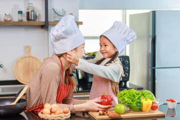 Millennial Asian young little cute girl chef daughter in apron standing smiling wearing adjusting white tall cook hat for mother ready for cooking vegetables food meal at counter in home kitchen