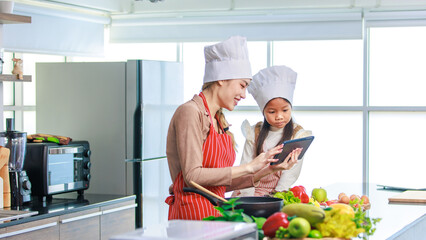 Asian young female chef housewife mother wears white tall cook hat and apron showing mockup glass touchscreen tablet computer screen to little cute girl chef learning cooking online at home kitchen