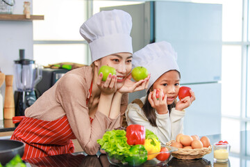 Asian young female chef housewife mother and little cute girl daughter wears white tall cook hat and apron smiling posing looking  at home kitchen.