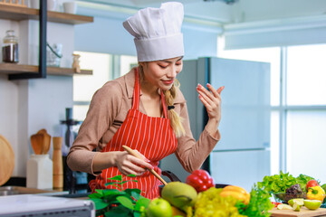 Millennial Asian young cheerful female housewife chef wearing white tall cook hat and apron standing smiling waving hand smelling tasty delicious meal while cooking in pan in full decorated kitchen