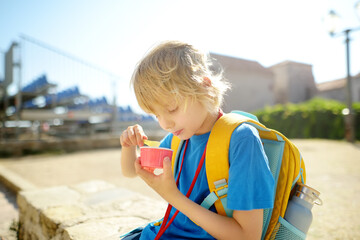 Happy little boy eating tasty ice cream in paper cup outdoors. School child have ice-cream snack on the go after school on sunny day. Kids love gelato