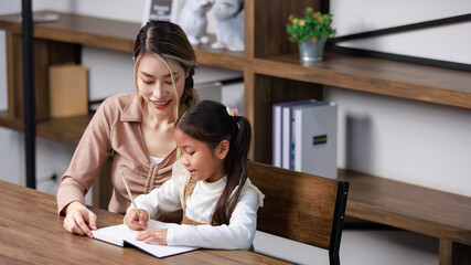 Asian young female housewife mother tutor teacher sitting smiling on table in living room at home teaching little cute kindergarten preschool girl daughter writing on book doing homework after school