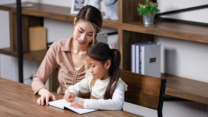 Asian young female housewife mother tutor teacher sitting smiling on table in living room at home teaching little cute kindergarten preschool girl daughter writing on book doing homework after school