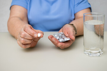 Senior woman with wrinkled old hands at the table holding one round medical tablet, painkiller or pill for treatment and water glass. Healthcare and medicine concept
