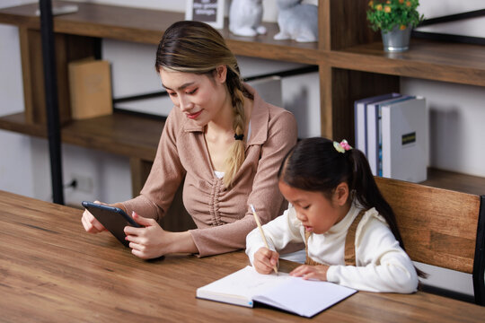 Asian Young Female Housewife Mother Tutor Teacher Sitting Smiling On Table In Living Room Holding Tablet Computer Pointing Teaching Little Cute Kindergarten Preschool Girl Daughter Doing Homework