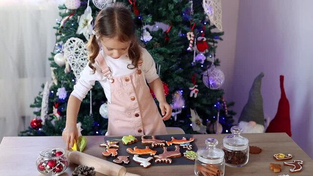Happy little girl coloring Christmas gingerbread cookies at home. Christmas decoration in the kitchen. Fir tree with fairy lights. The concept of the New Year and Christmas. Holiday atmosphere