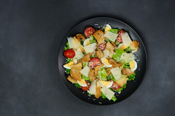 Top view of a Caesar Salad with chicken, lettuce leaves, cherry tomatoes, grated parmesan in a black plate against a Black Background