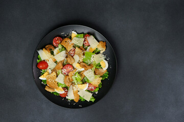 Top view of a Caesar Salad with chicken, lettuce leaves, cherry tomatoes, grated parmesan in a black plate against a Black Background