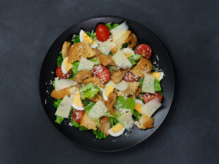 Top view of a Caesar Salad with chicken, lettuce leaves, cherry tomatoes, grated parmesan in a black plate against a Black Background