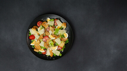 Caesar Salad with chicken, lettuce leaves, cherry tomatoes, grated parmesan in a black plate against a Black Background
