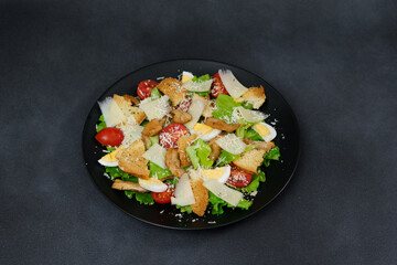 Caesar Salad with chicken, lettuce leaves, cherry tomatoes, grated parmesan in a black plate against a Black Background
