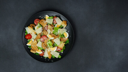 Top view of a Caesar Salad with chicken, lettuce leaves, cherry tomatoes, grated parmesan in a black plate against a Black Background