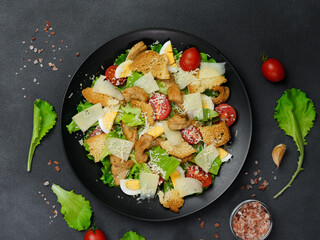 Top view of a Caesar Salad with chicken, lettuce leaves, cherry tomatoes, grated parmesan in a black plate against a Black Background