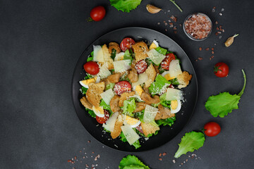 Top view of a Caesar Salad with chicken, lettuce leaves, cherry tomatoes, grated parmesan in a black plate against a Black Background