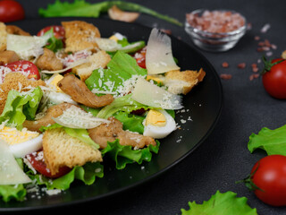 Close-up of a Caesar Salad with chicken, lettuce leaves, cherry tomatoes, grated parmesan in a black plate against a Black Background
