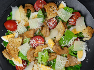 Close-up of a Caesar Salad with chicken, lettuce leaves, cherry tomatoes, grated parmesan in a black plate against a Black Background
