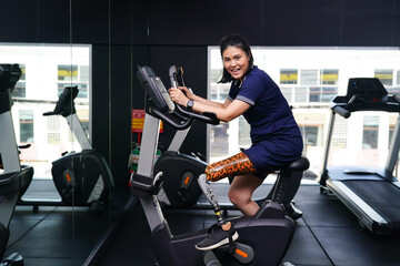 Young female with one prosthetic leg with exercising with a spinning bike in the gym to practice...